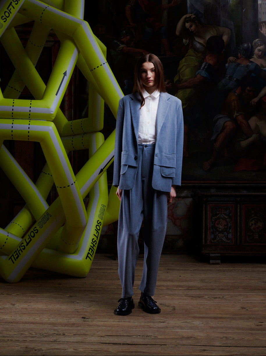 Model wearing grey Henrik Vibskov pants with drop crotch, styled with blazer in an industrial setting with green structure in background