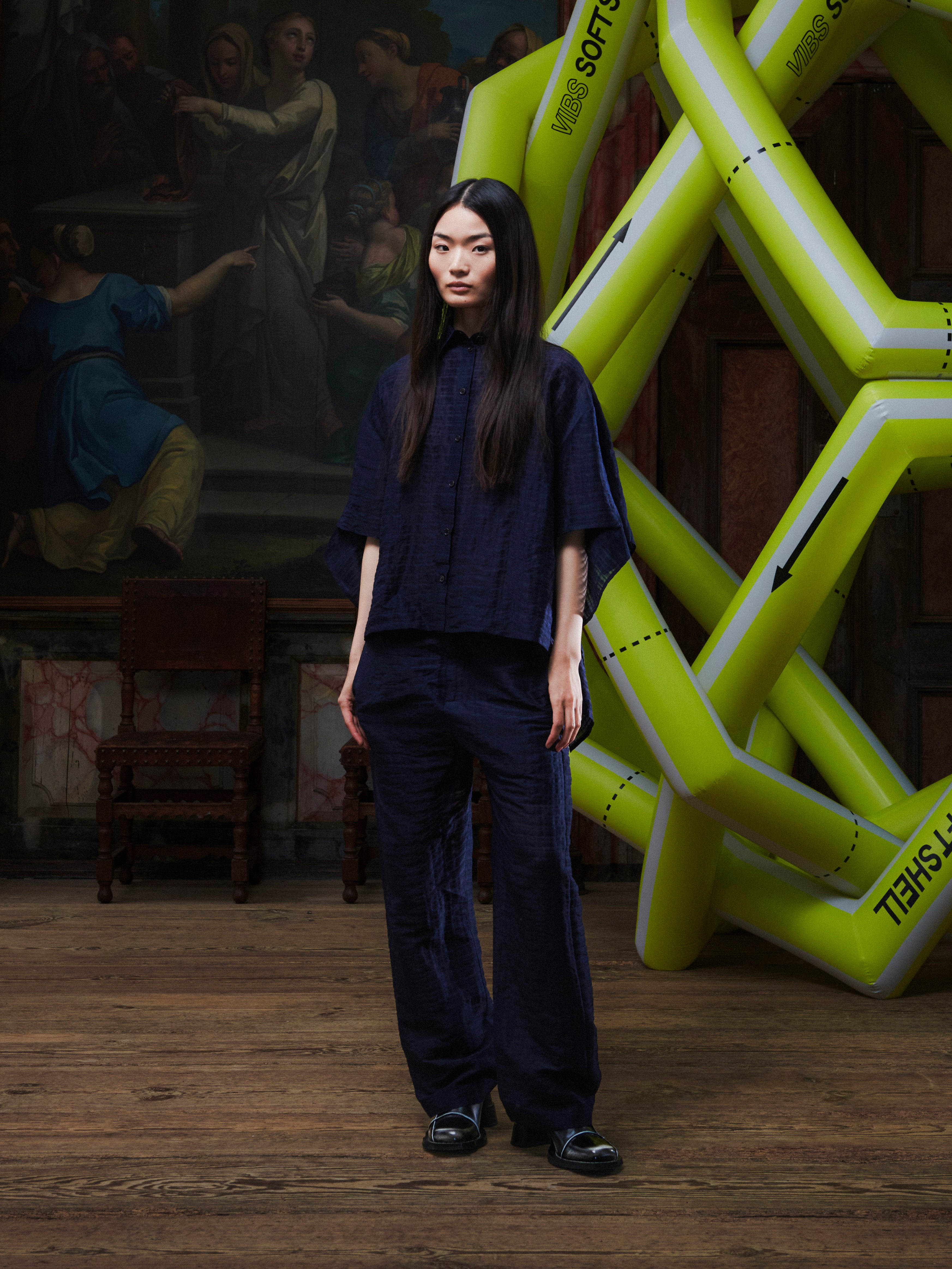 Front view of the female model wearing the Artist Pants in navy from Henrik Vibskov Spring/Summer 2026 Studio Collection, showing the unisex straight-leg fit, front button and zipper closure, and side pockets.