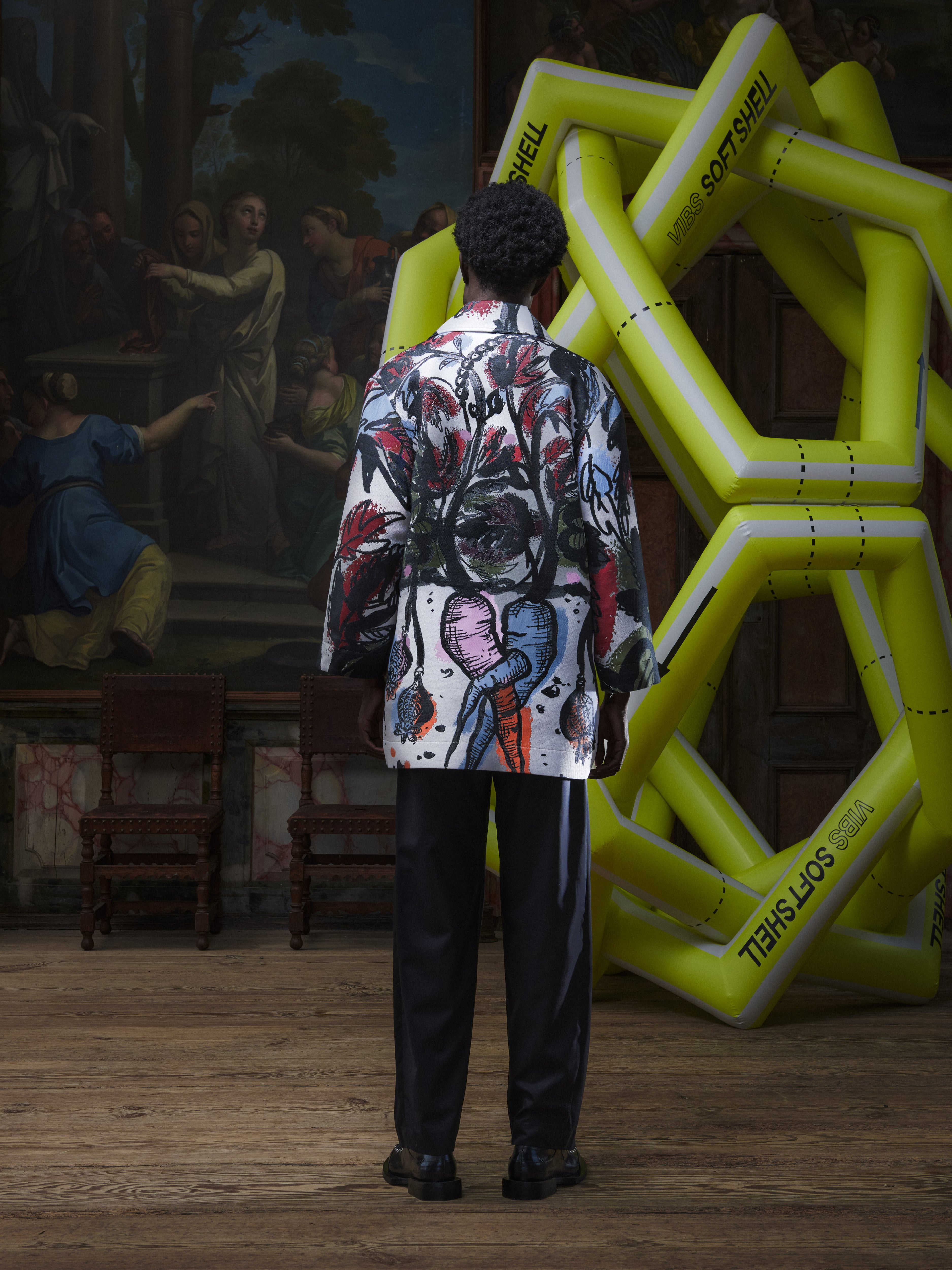 Back view of the male model wearing the Care Coat in Carrots from Henrik Vibskov Spring/Summer 2026 Studio Collection, highlighting the full carrot pattern across the back and straight hemline.
