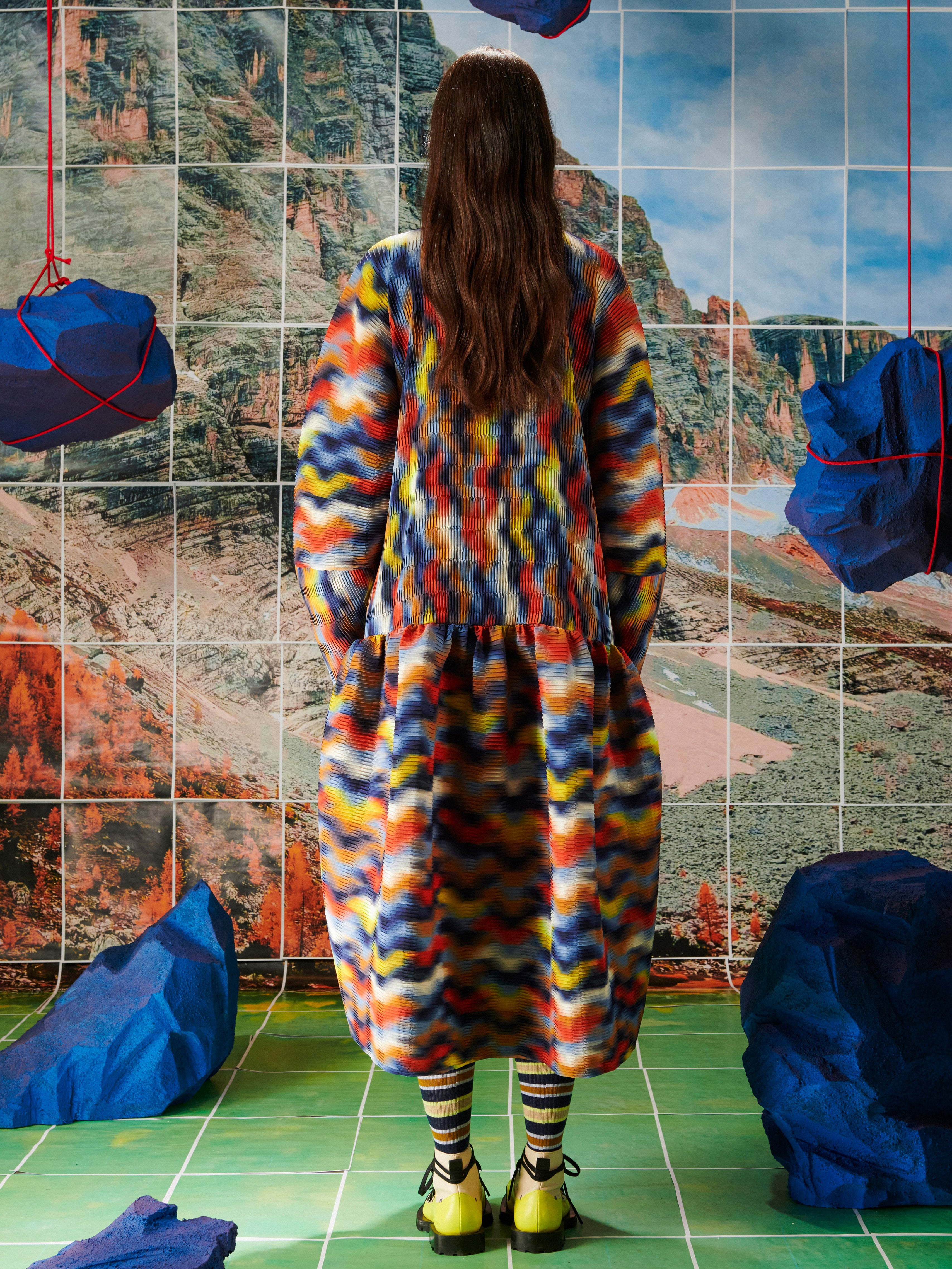Person wearing a colorful dress from Henrik Vibskov standing in front of a tiled wall with abstract patterns