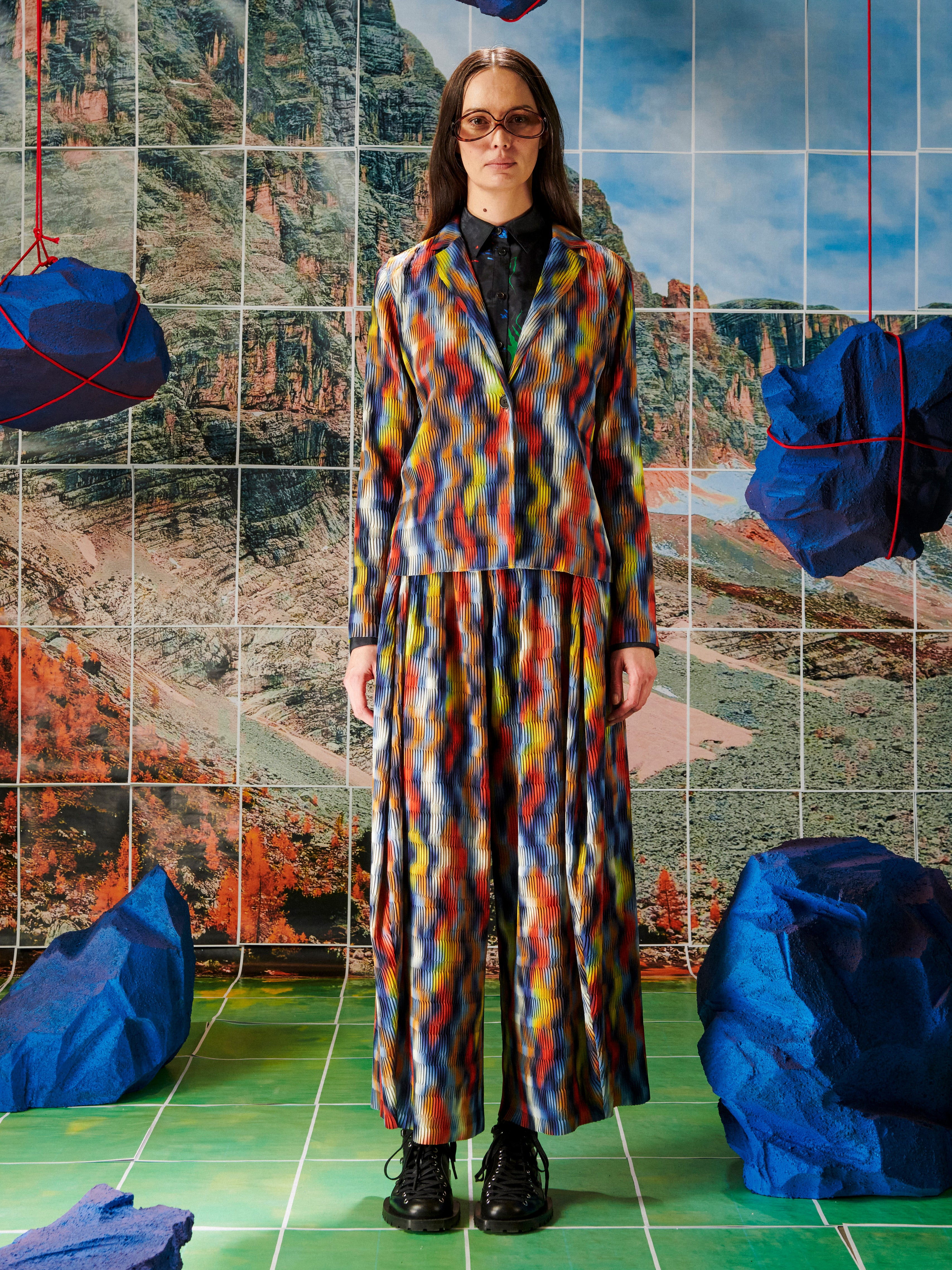 Person wearing a colorful outfit standing in front of a tiled wall with abstract patterns.
