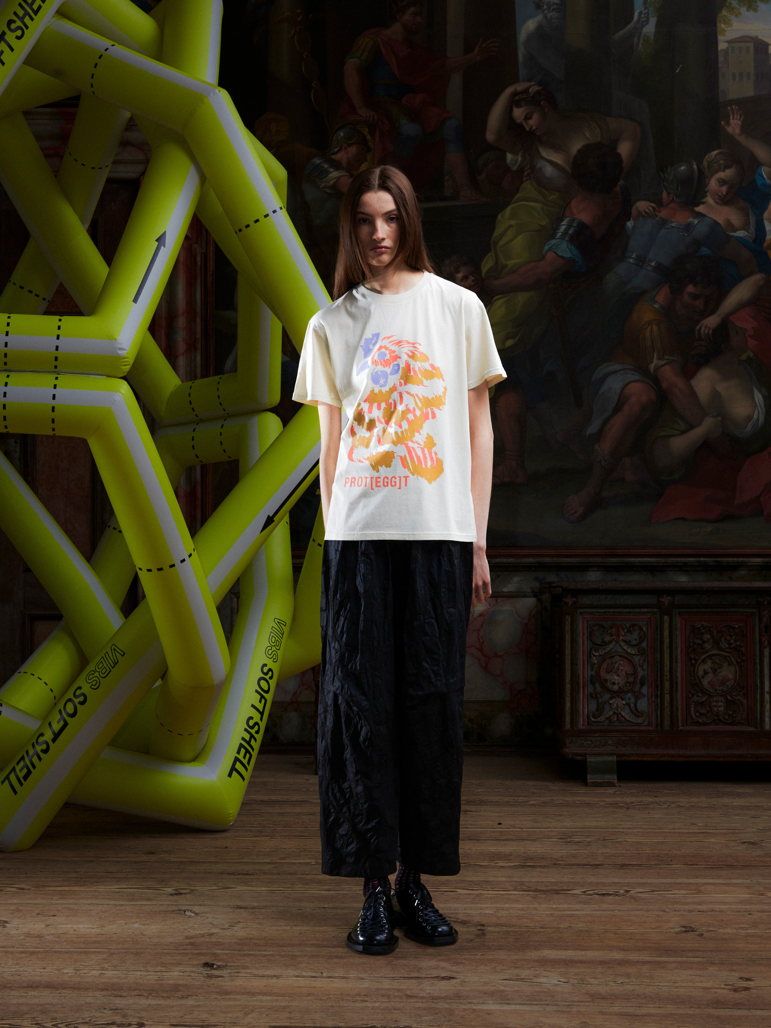 Female model wearing the off-white t-shirt with an orange rooster illustration by Henrik Vibskov in a setting with an inflatable structure