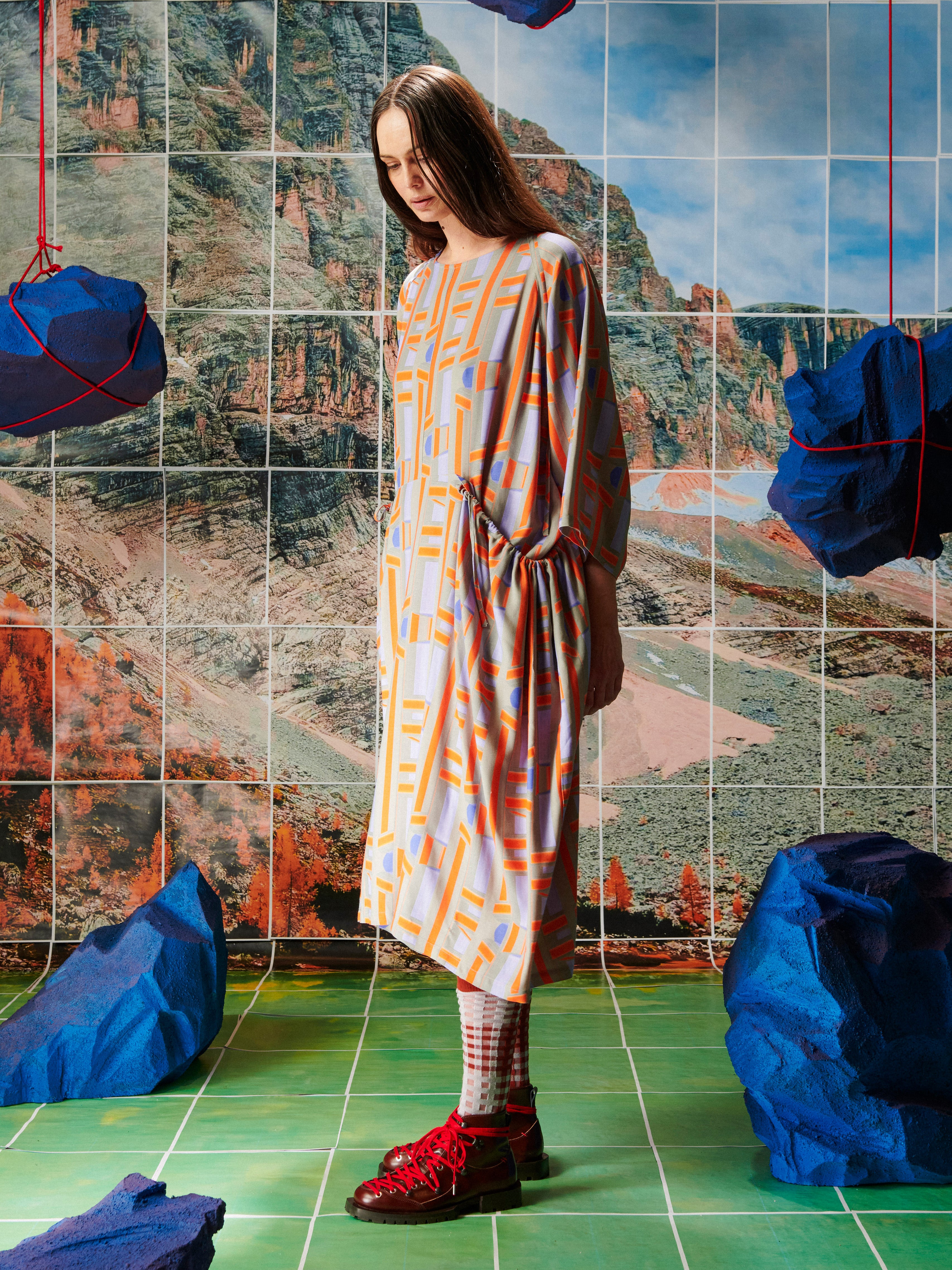 Woman in a patterned dress by Henrik Vibskov standing in front of a tiled wall with abstract designs.