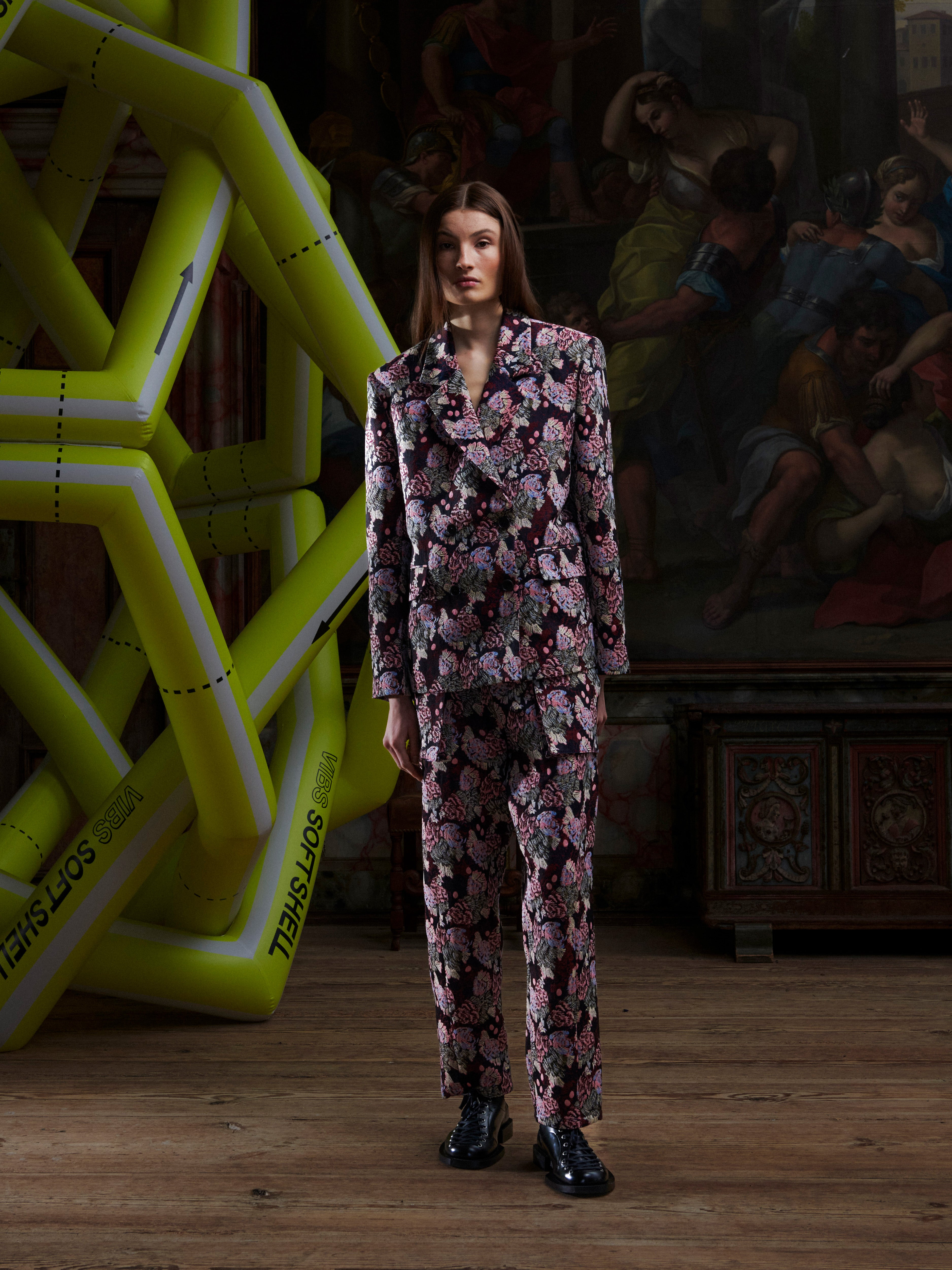 Female model wearing a patterned suit by Henrik Vibskov, standing in front of a large inflatable sculpture with a painting in the background.