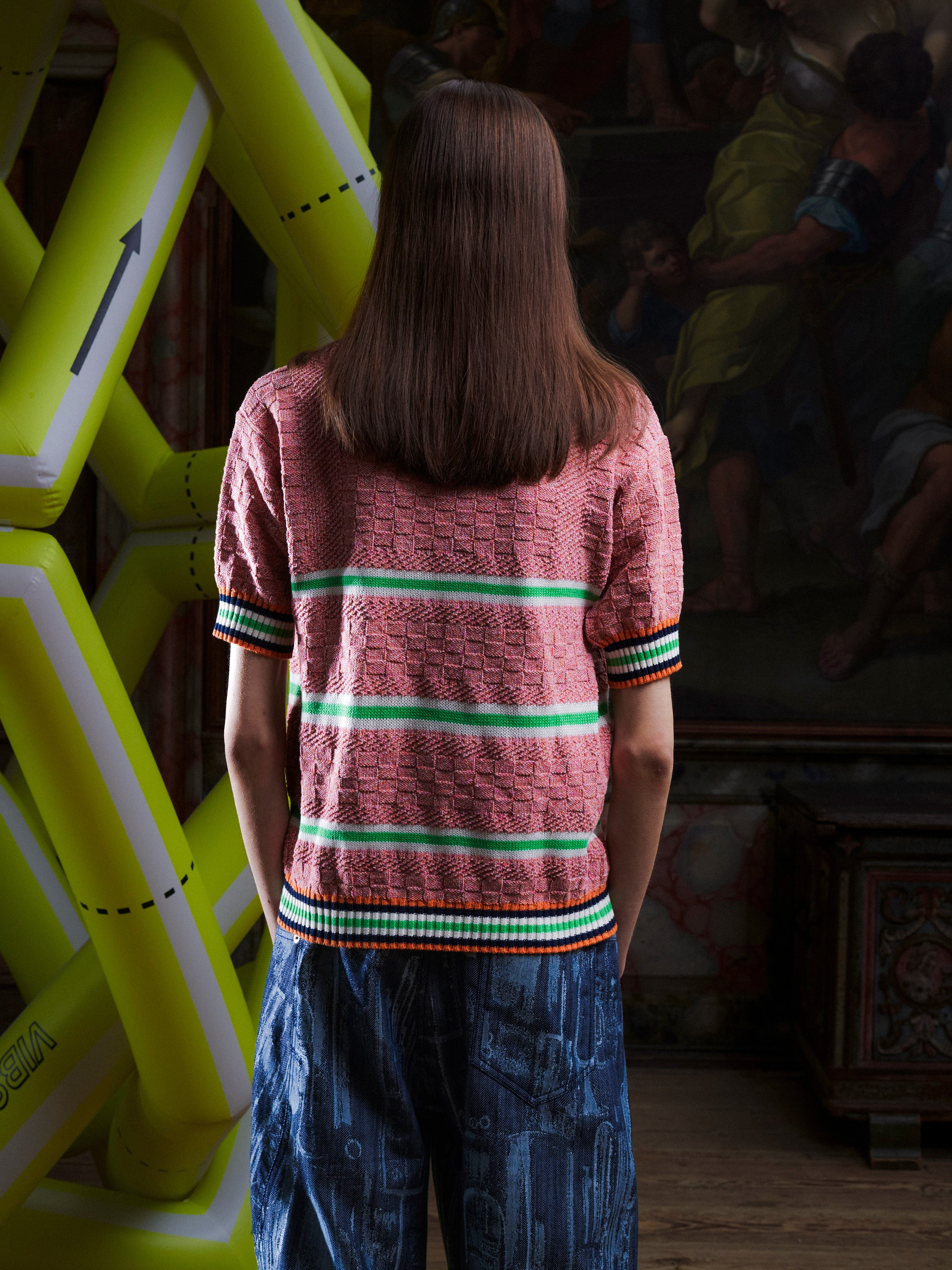 Back view of woman wearing the pink-green knitted top with stripe details from Henrik Vibskov with a yellow inflatable sculpture in the background.