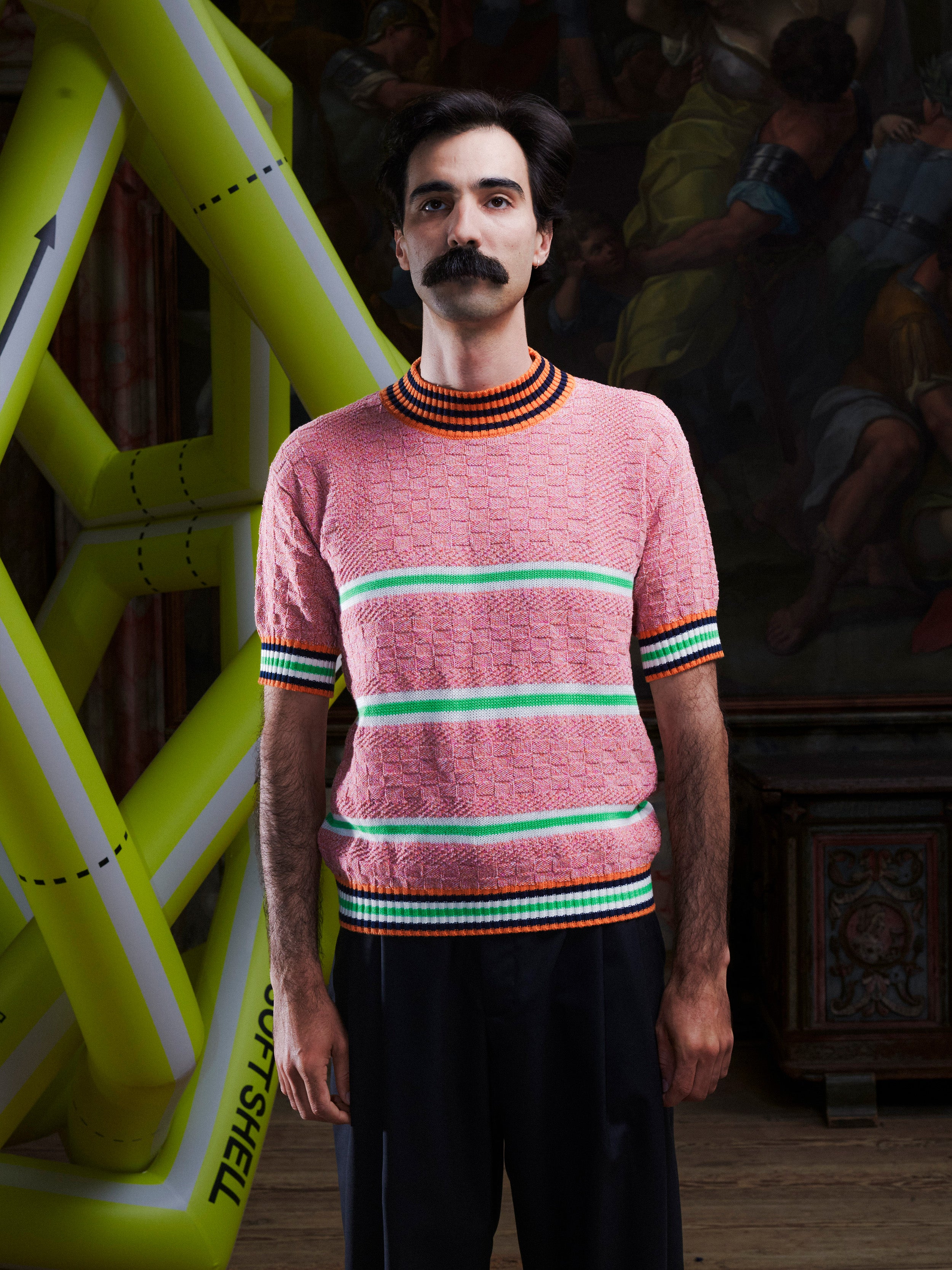 Man wearing the pink-green knitted top with stripe details from Henrik Vibskov with a yellow inflatable sculpture in the background.