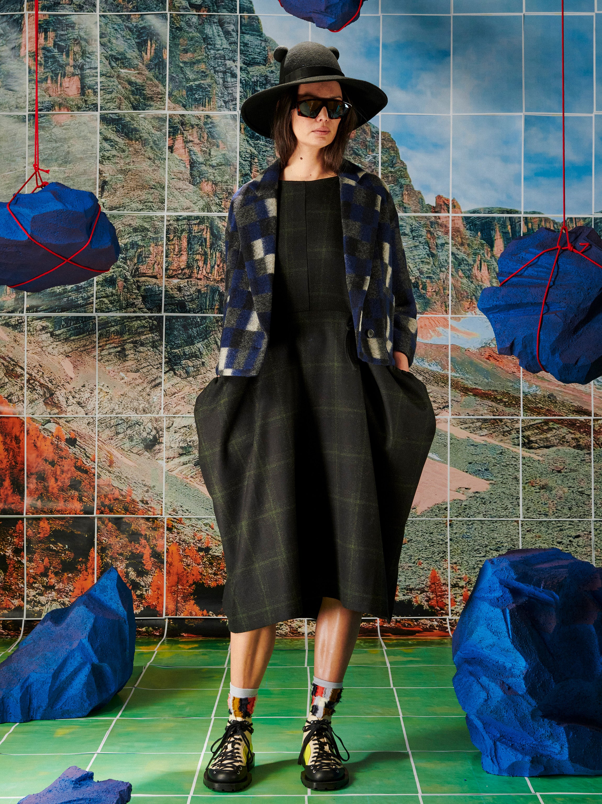Person wearing a plaid jacket and black hat in front of a tiled wall with blue and red abstract art.