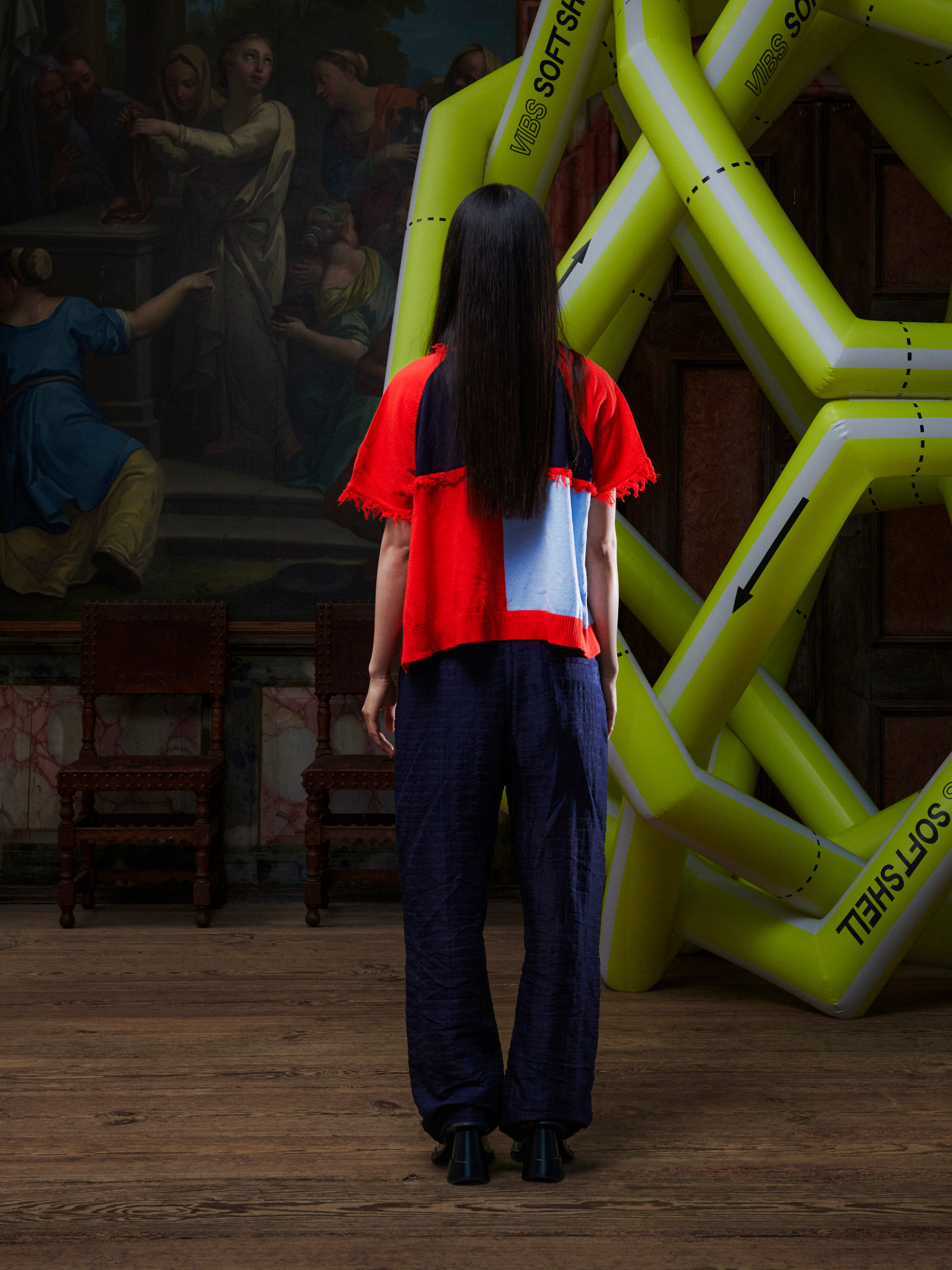 Back profile of person wearing a bright red knitted short sleeve shirt standing in front of a large inflatable structure and classical painting.