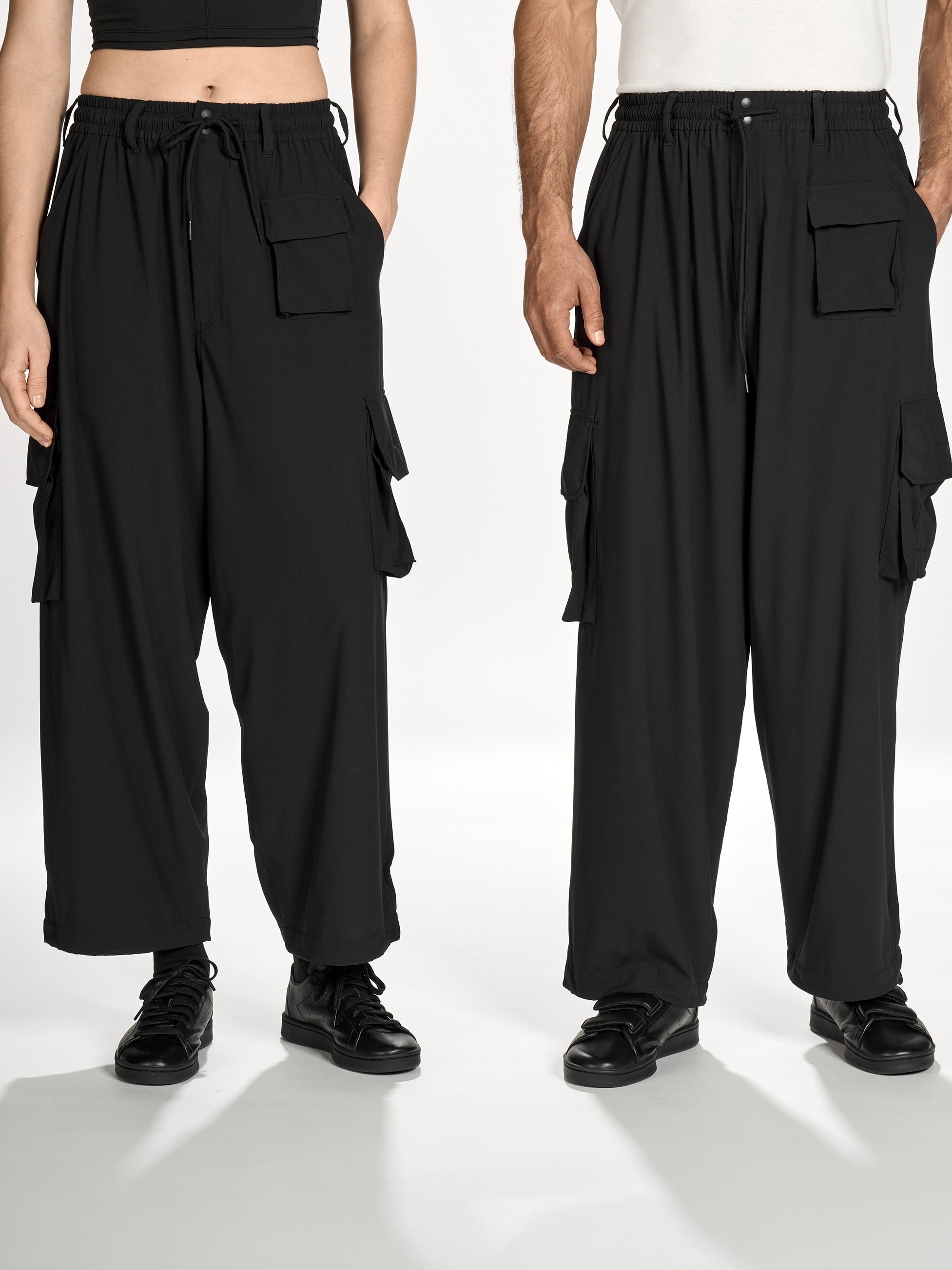 Side view of female and male models wearing Y-3 Utility Straight Tracksuit Pants in Black, highlighting the straight-leg silhouette and drape of the plain weave fabric.