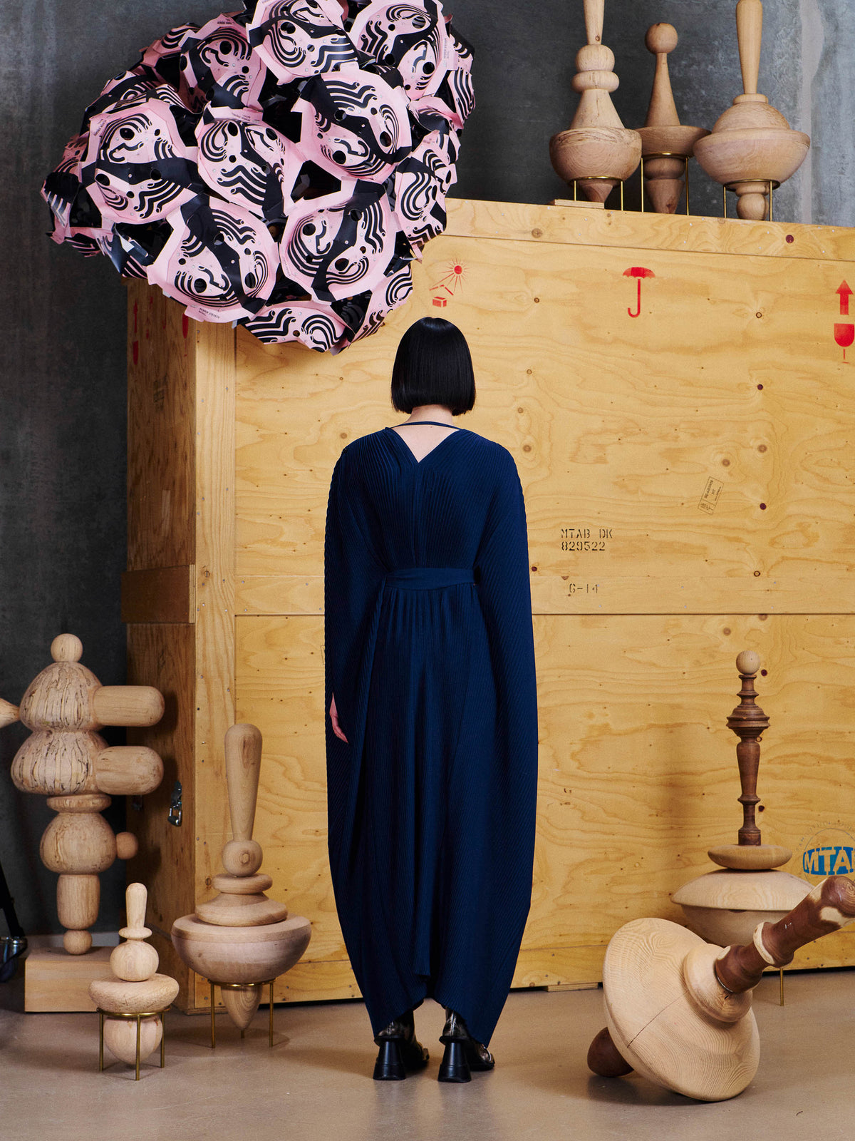 Back view of model wearing Henrik Vibskov Cherry Plisse Kaftan in Dress Blues, showing the V-shaped back neckline, tied waist and long pleated silhouette.