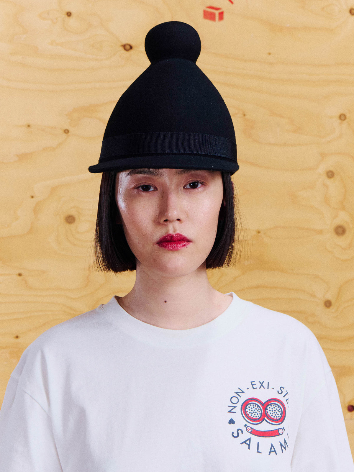 Model wearing the Henrik Vibskov Jockey Cap in Black, showing the rounded crown, top pom-pom, curved brim and grosgrain band.
