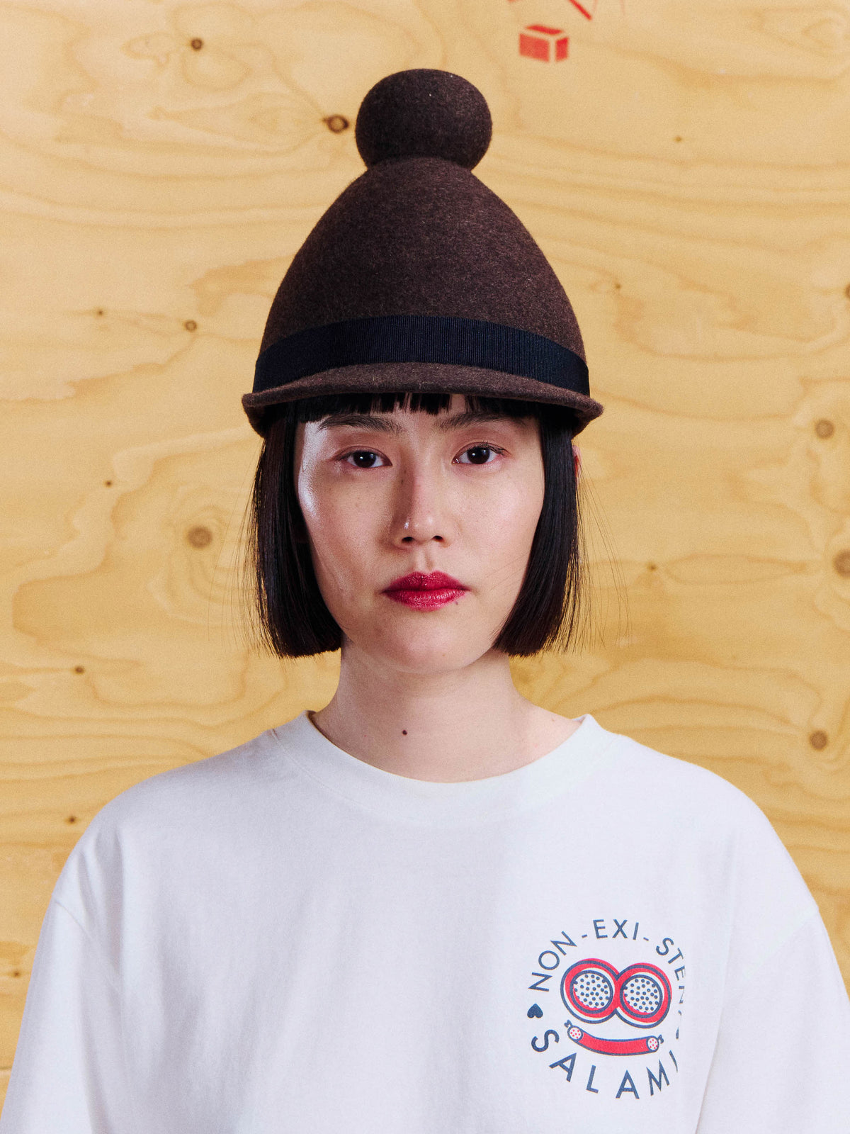 Model wearing the Henrik Vibskov Jockey Cap in Brown, showing the rounded crown, top pom-pom, curved brim and contrast band.