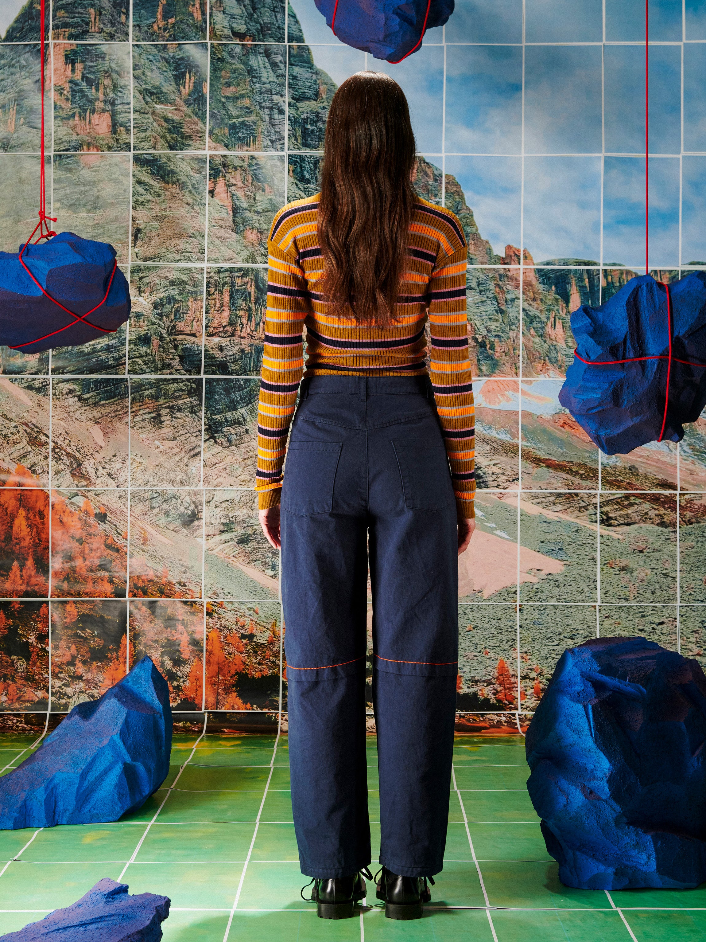 Person wearing a striped sweater and dark pants standing in front of a tiled wall with mountain scenes.