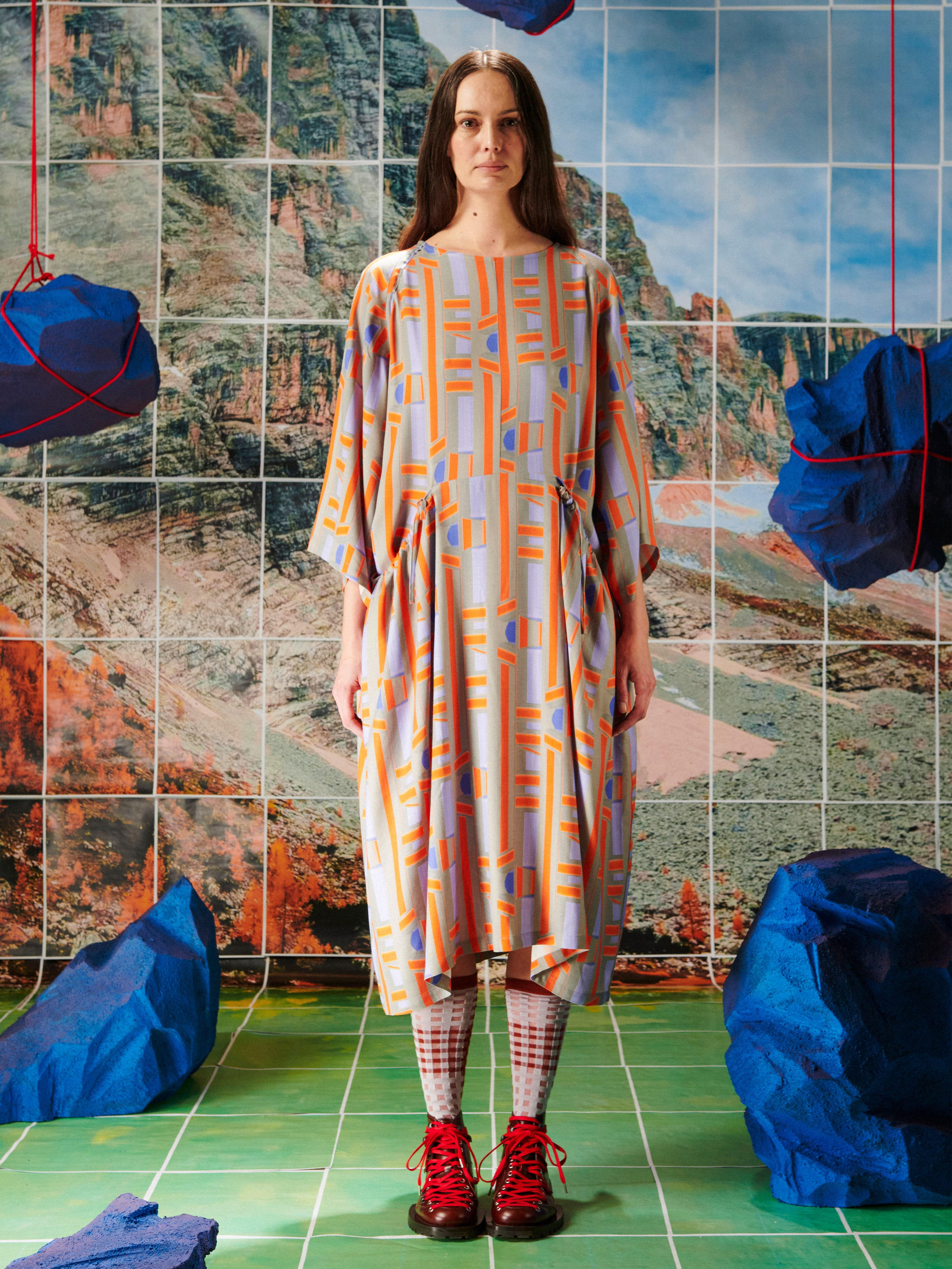 Woman in a patterned dress by Henrik Vibskov standing in front of a tiled wall with abstract designs.