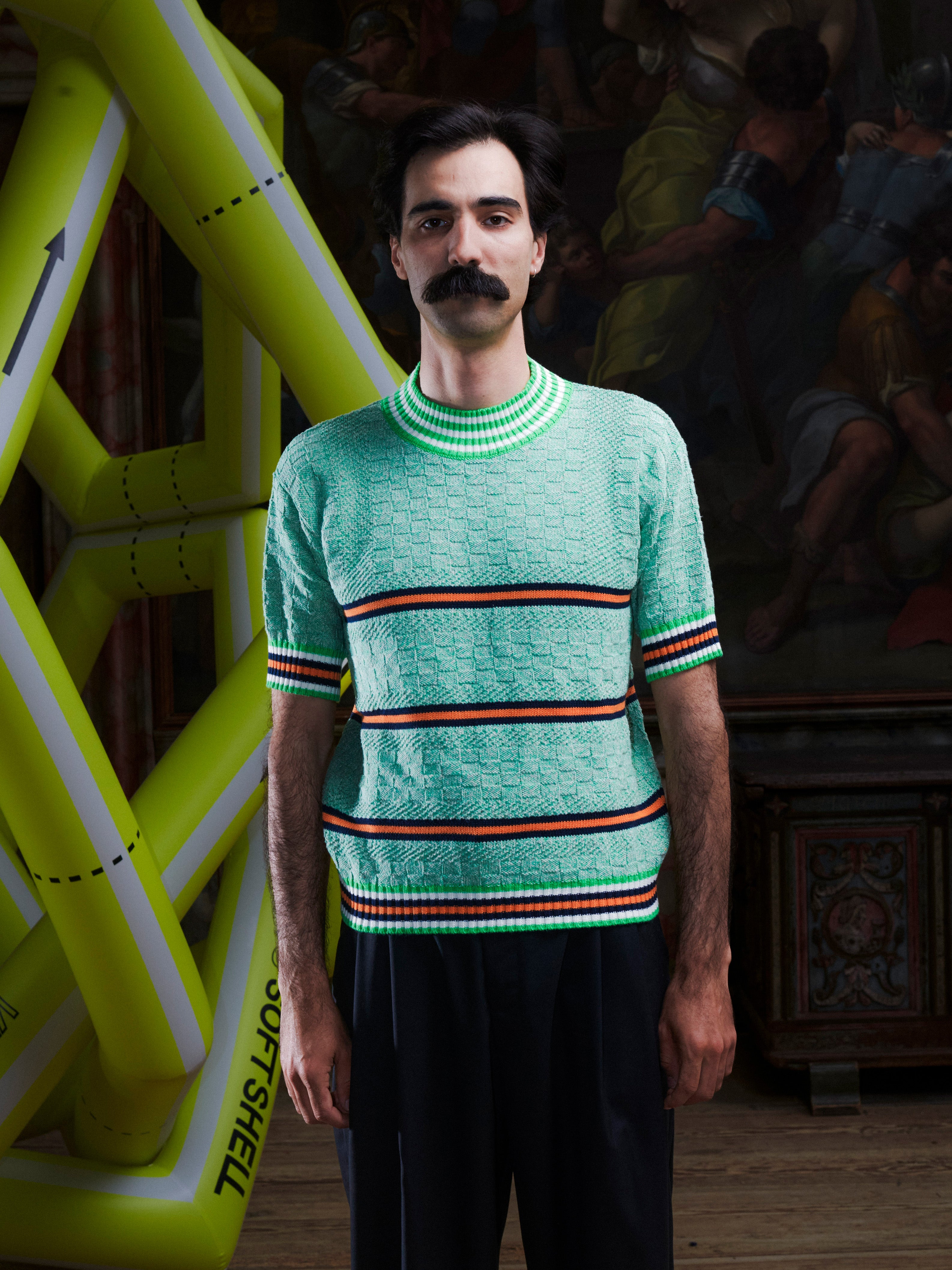 Man wearing the mint green knitted top with stripe details from Henrik Vibskov with a yellow inflatable sculpture in the background.