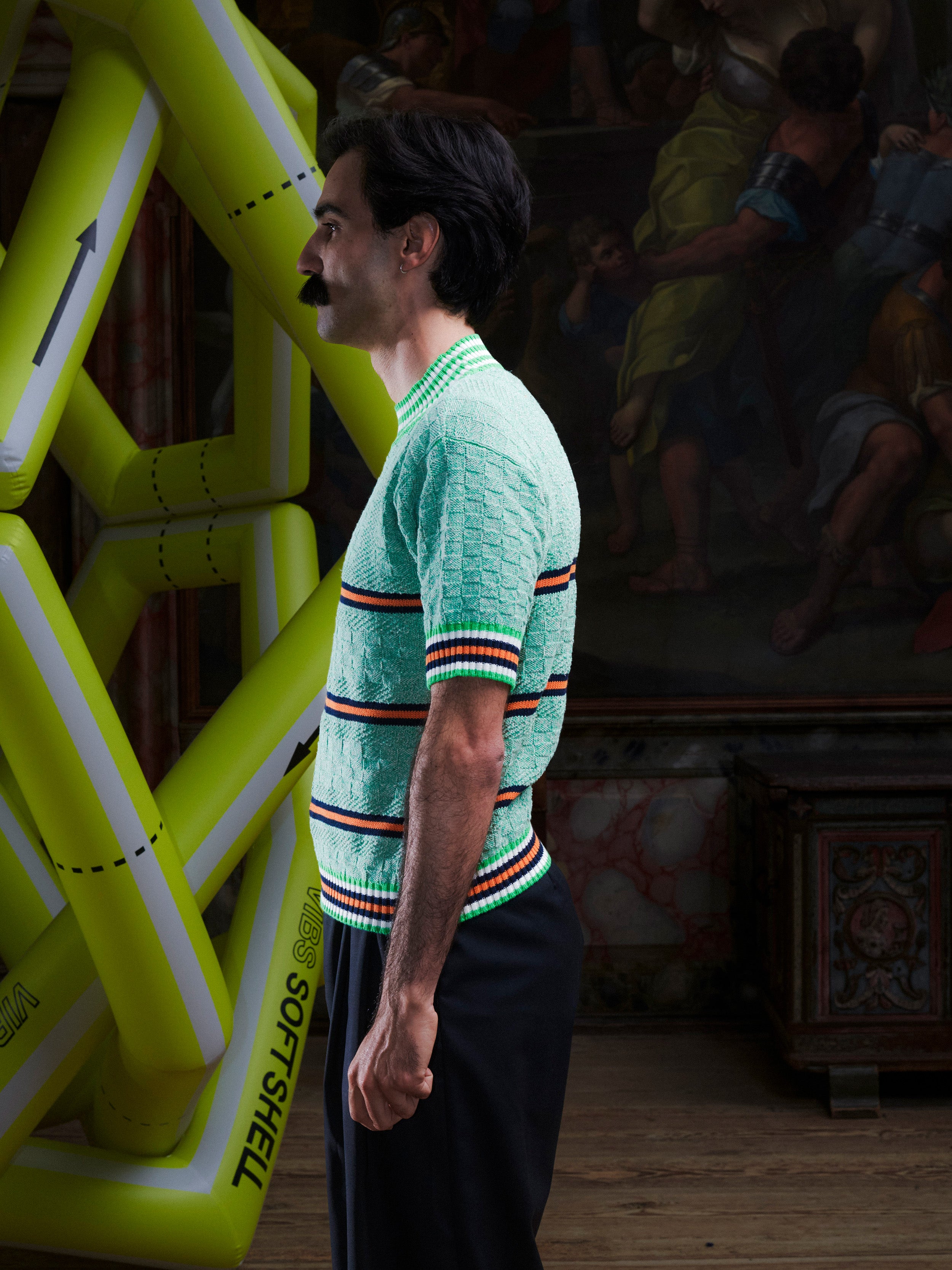 Side profile of man wearing the mint green knitted top with stripe details from Henrik Vibskov with a yellow inflatable sculpture in the background.