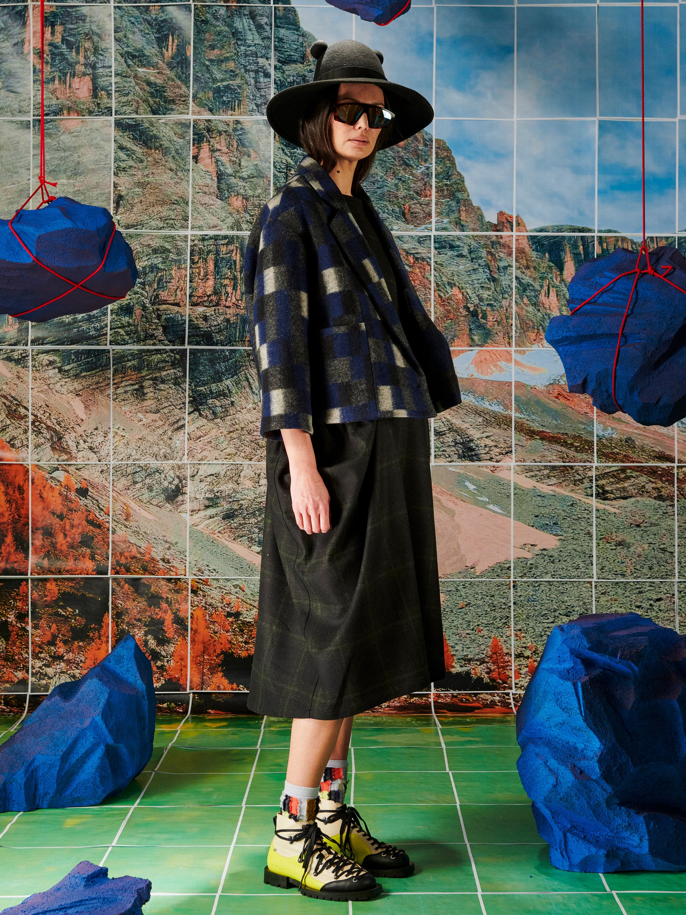 Person wearing a plaid jacket and black hat in front of a tiled wall with blue and red abstract art.