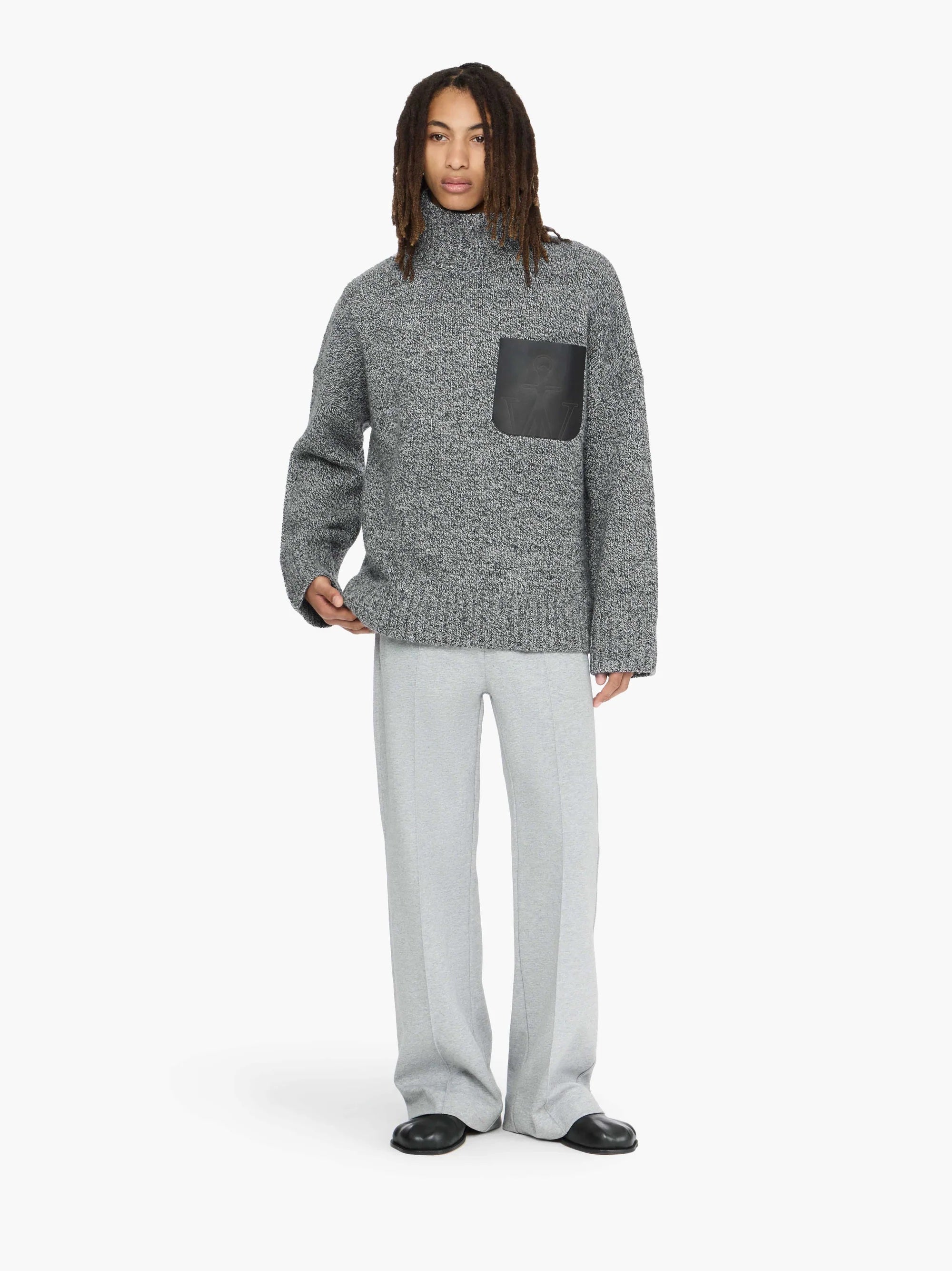 Person wearing a gray sweater with a black pocket and light gray pants on a white background
