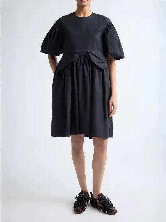 Model wearing the Simone Rocha Mini Puff Sleeve Gathered Bow Smock Dress with puff sleeves, gathered skirt and sculptural front bow detail.